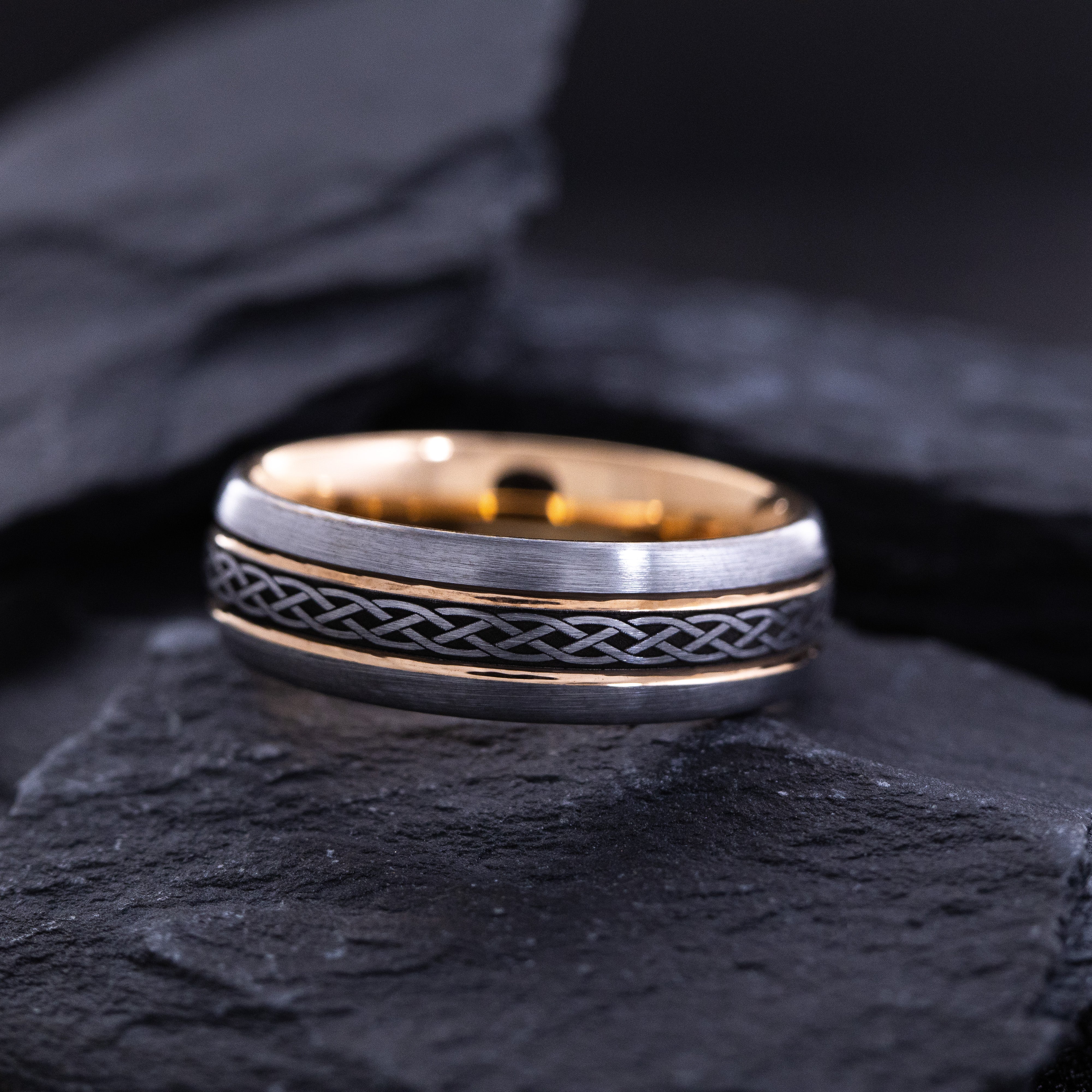 Two rose gold and silver tungsten rings with intricate designs on a dark stone surface